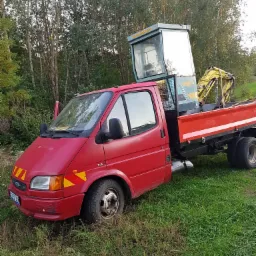 Czerwony samochód dostawczy z otwartymi drzwiami kierowcy, przewożący małą żółtą koparkę na platformie, stojący na zarośniętym terenie na tle drzew.