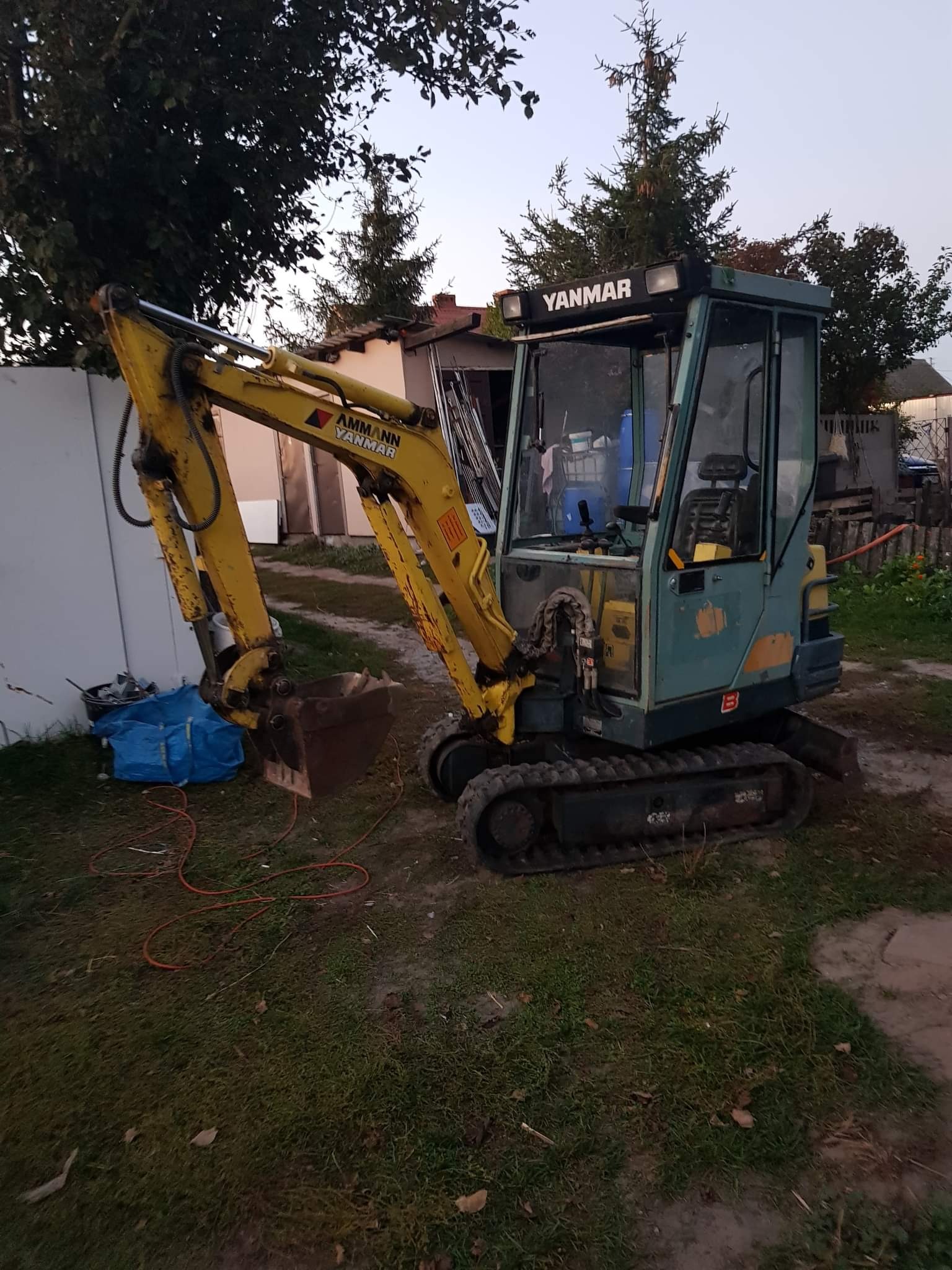 Żółto-zielona mini koparka Yanmar na trawiastym terenie z widocznymi śladami użytkowania, obok białego ogrodzenia i pod drzewami.