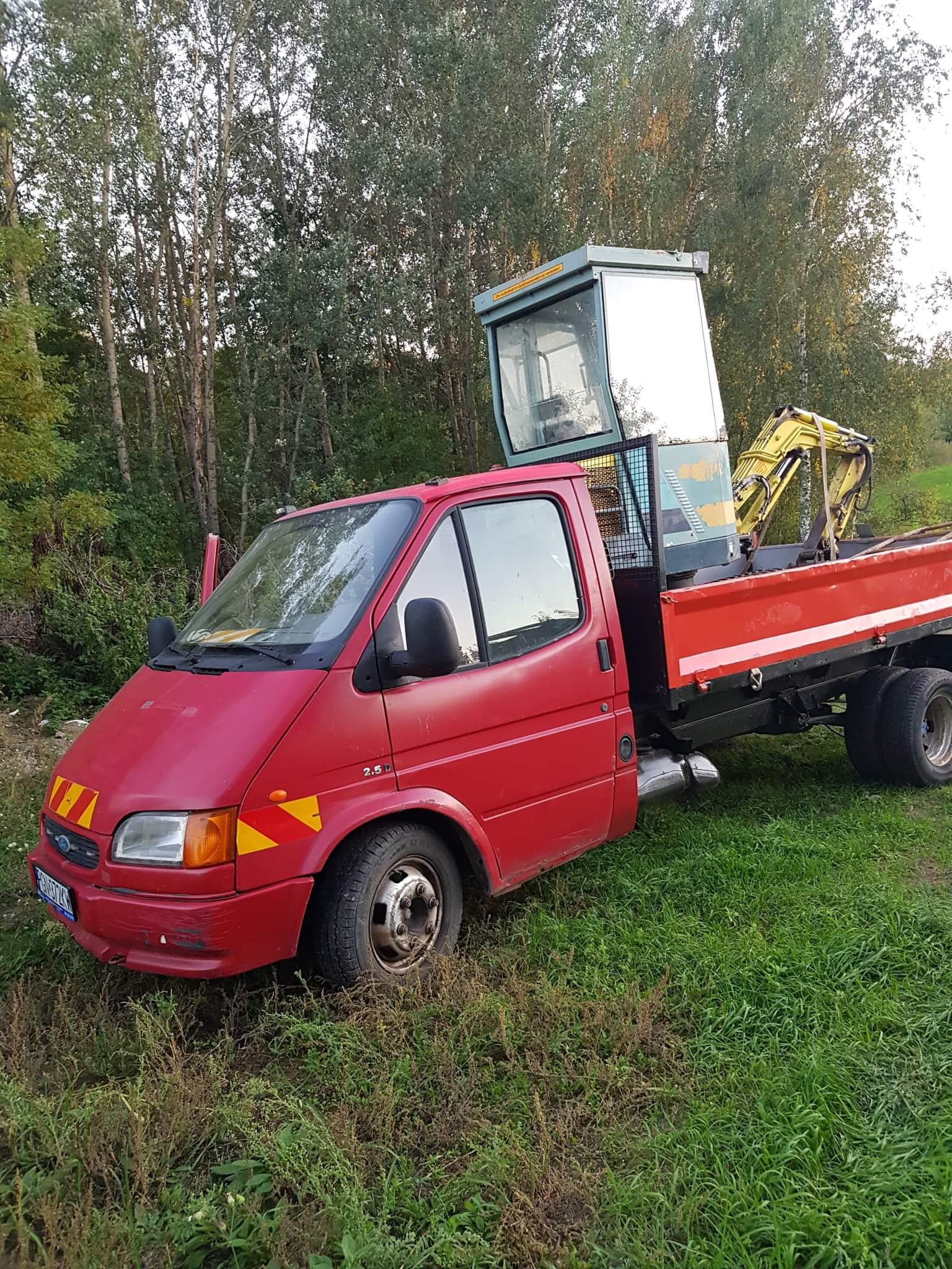 Czerwony samochód dostawczy z otwartymi drzwiami kierowcy, przewożący małą żółtą koparkę na platformie, stojący na zarośniętym terenie na tle drzew.