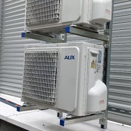 Lukwent - Dwa białe klimatyzatory AUX zamontowane jeden nad drugim na metalowej konstrukcji wsporczej, na tle blaszanej ściany z poziomymi przetłoczeniami.