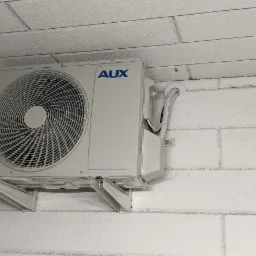 Biała jednostka zewnętrzna klimatyzacji marki AUX zamontowana na białej, ceglanej ścianie wewnętrznej, widoczne przewody w białej maskownicy.