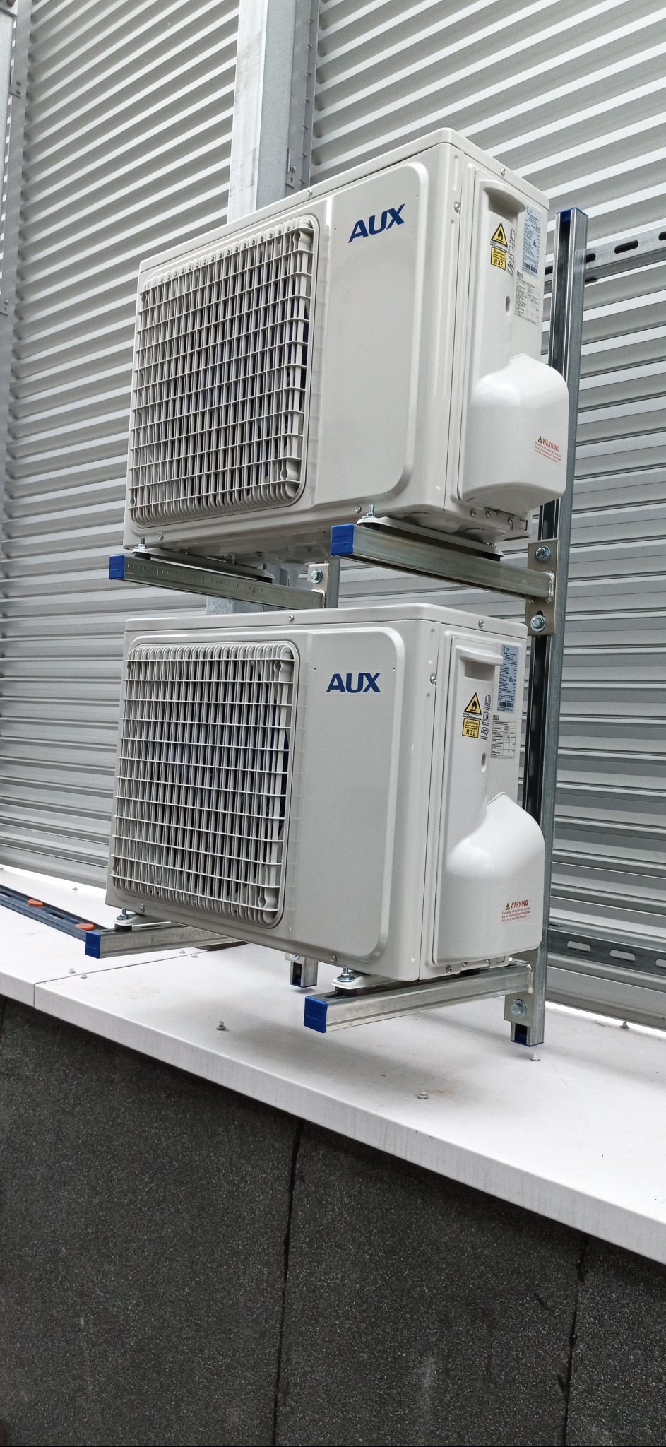Dwa białe klimatyzatory AUX zamontowane jeden nad drugim na metalowej konstrukcji wsporczej, na tle blaszanej ściany z poziomymi przetłoczeniami.