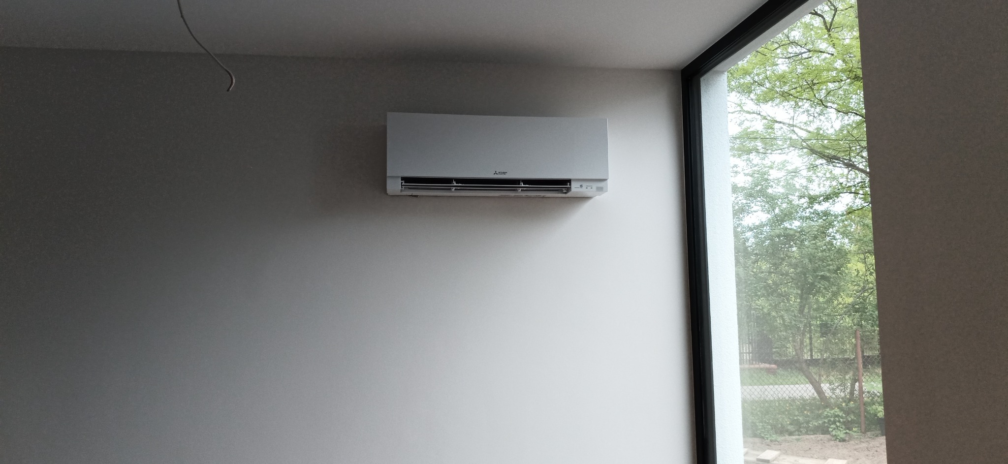 Biały klimatyzator Mitsubishi zamontowany na jasnej ścianie w minimalistycznym wnętrzu, widok na zieleń za oknem z czarną ramą.
