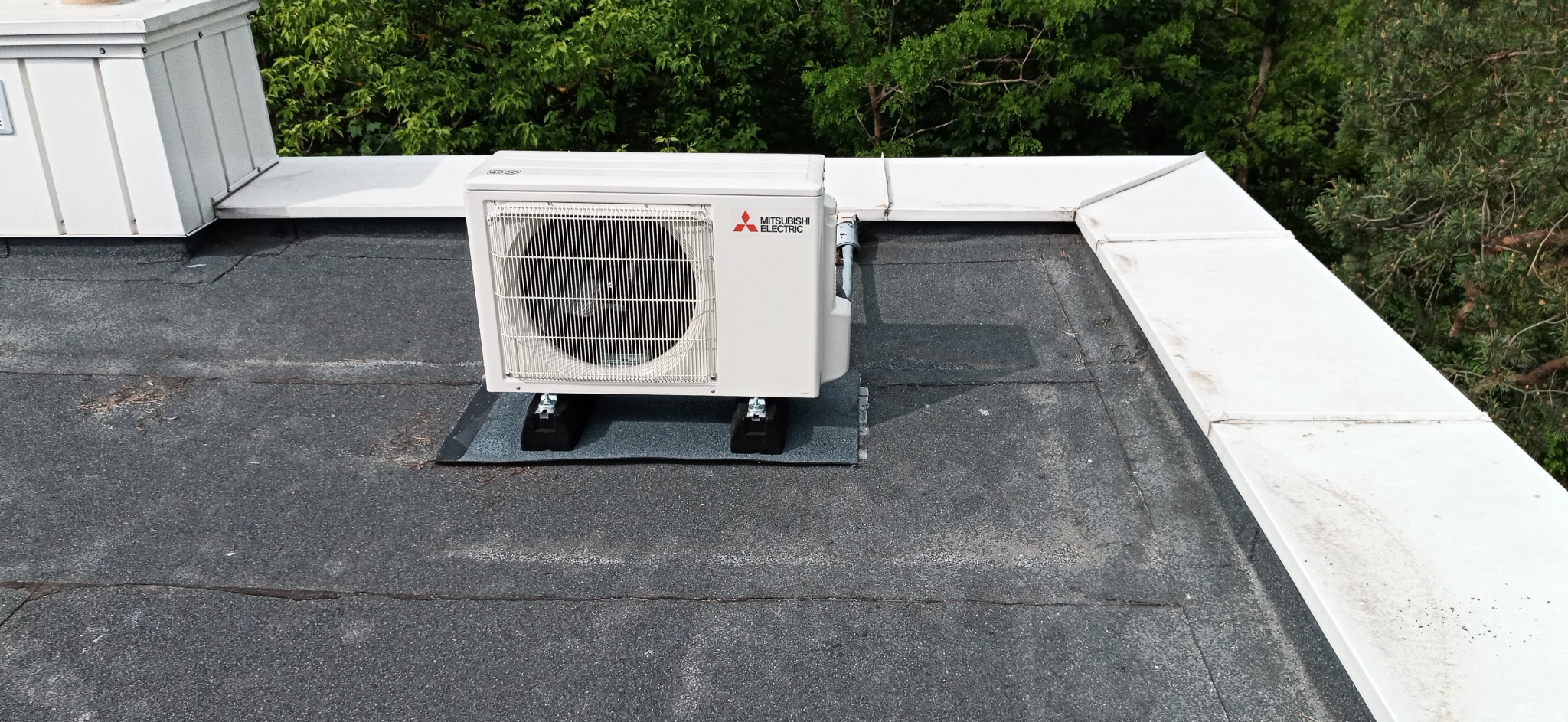 Biała jednostka zewnętrzna klimatyzatora Mitsubishi Electric, zamontowana na płaskim, bitumicznym dachu budynku, na gumowych podkładkach antywibracyjnych.