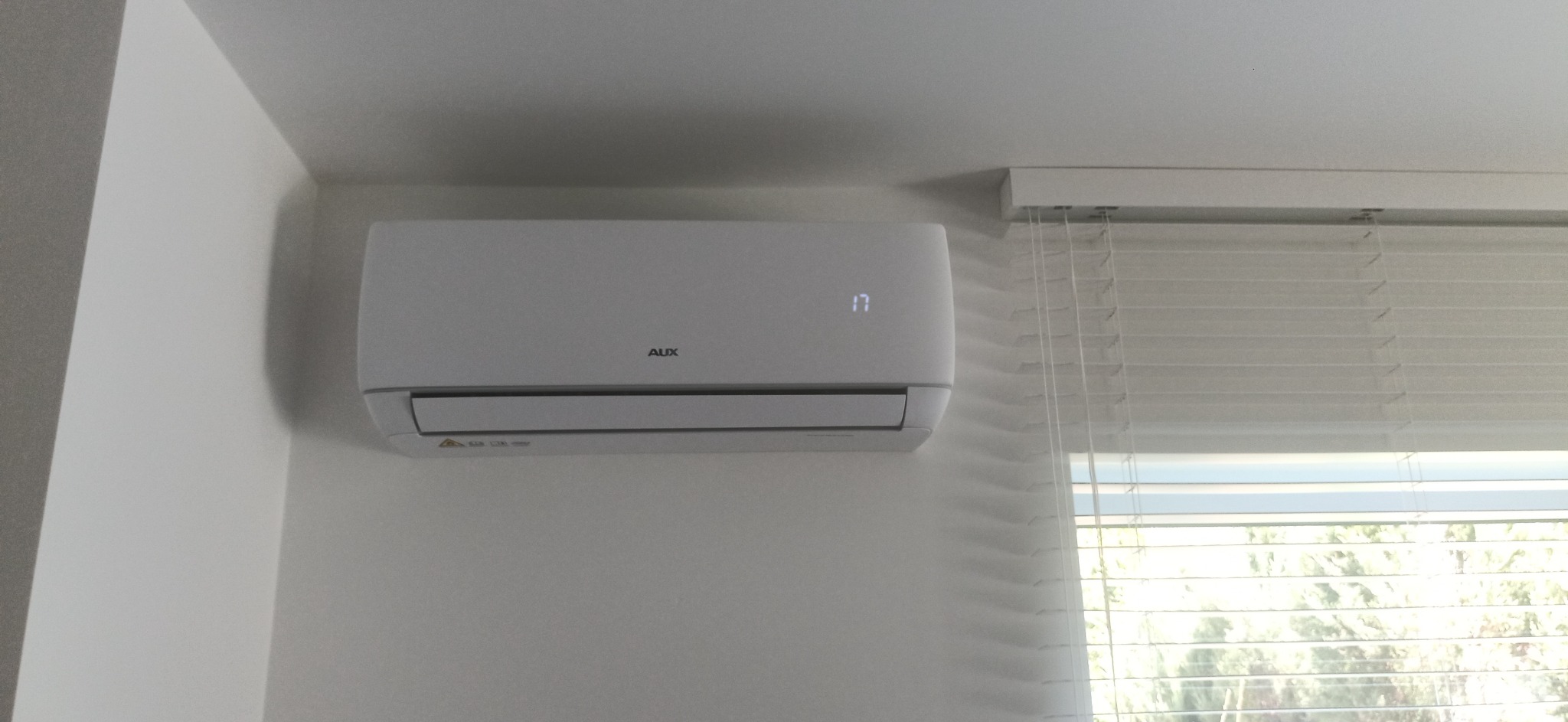 Biały klimatyzator marki AUX zamontowany na ścianie w pomieszczeniu, widoczny wyświetlacz z temperaturą 17 stopni Celsjusza, obok okno z żaluzjami, neutralne tło ścian i sufitu.