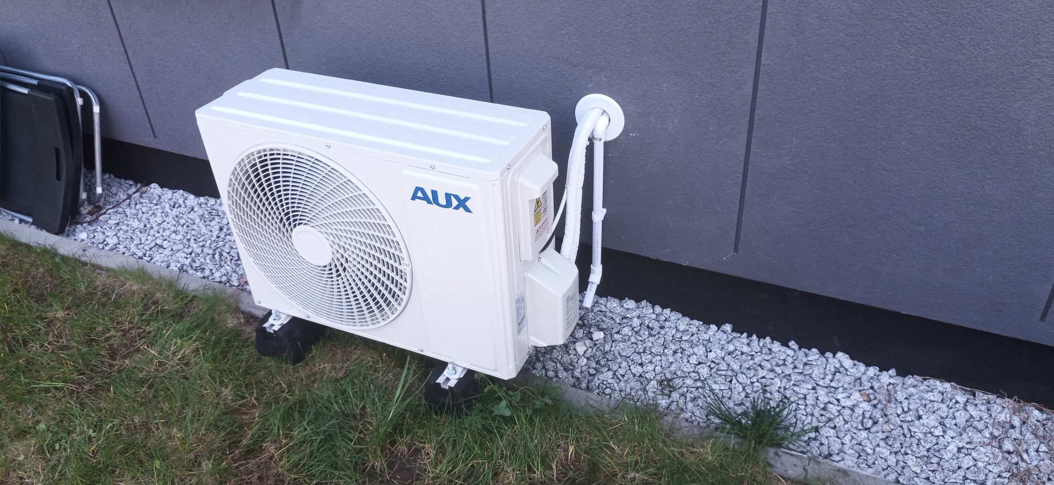 Zewnętrzna jednostka klimatyzacji marki AUX, zamontowana na gumowych podkładkach na trawie, z widocznymi rurami przyłączeniowymi wchodzącymi w ścianę budynku.