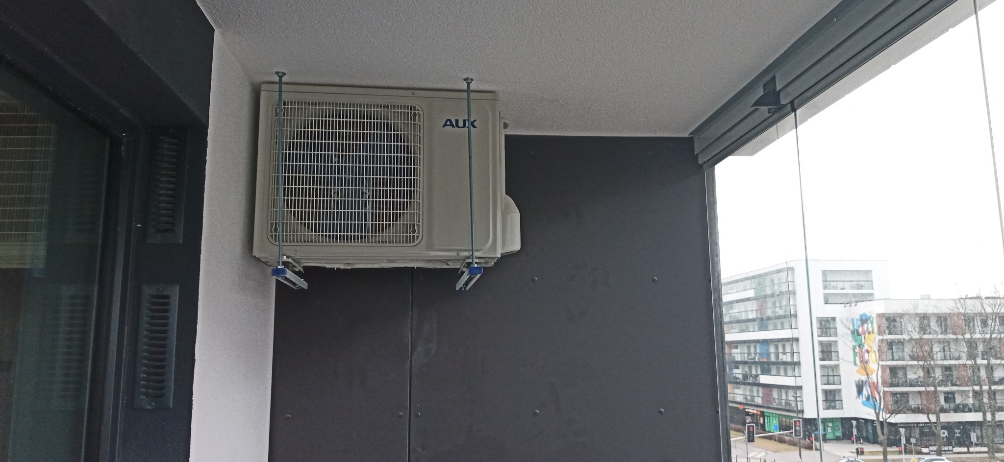 Klimatyzator marki AUX zamontowany na balkonie w bloku mieszkalnym, widoczne stalowe wsporniki i fragment panoramy miasta za szybą.