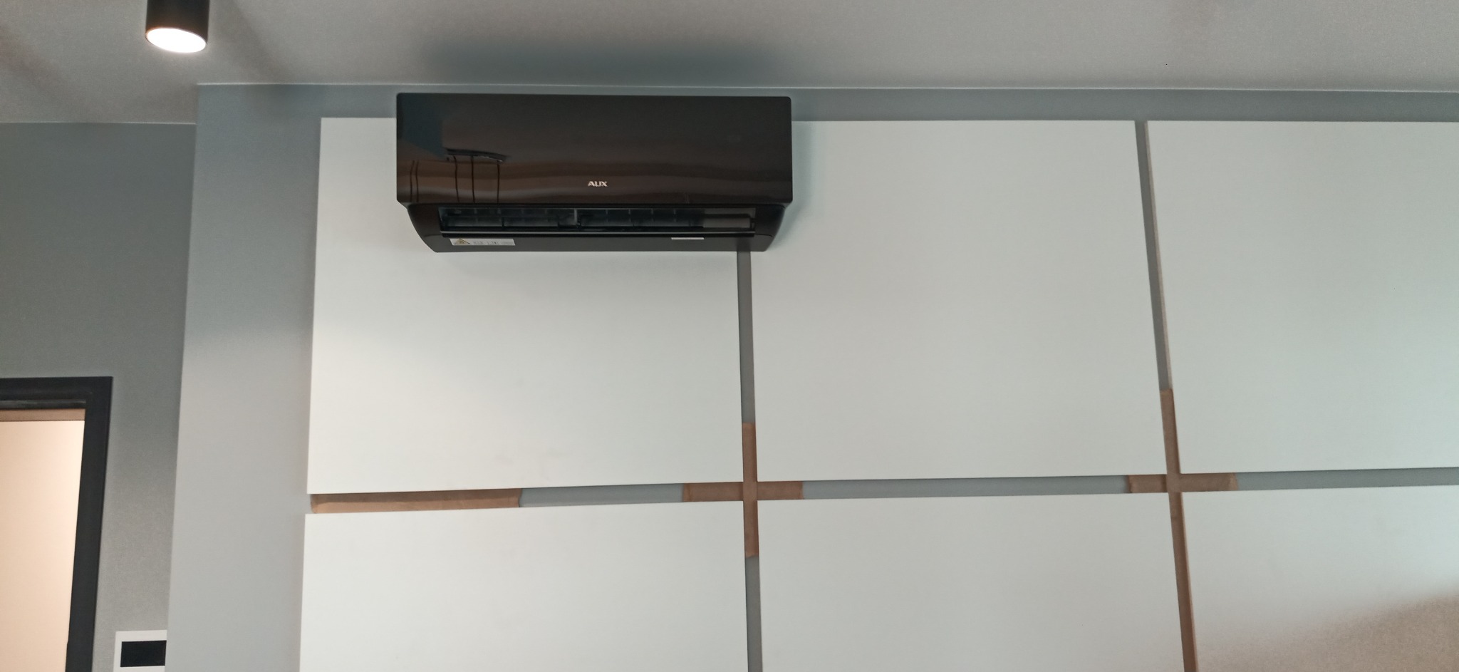 Czarny klimatyzator marki AUX zamontowany na białej, panelowej ścianie z elementami dekoracyjnymi w odcieniach drewna, widok z dołu.