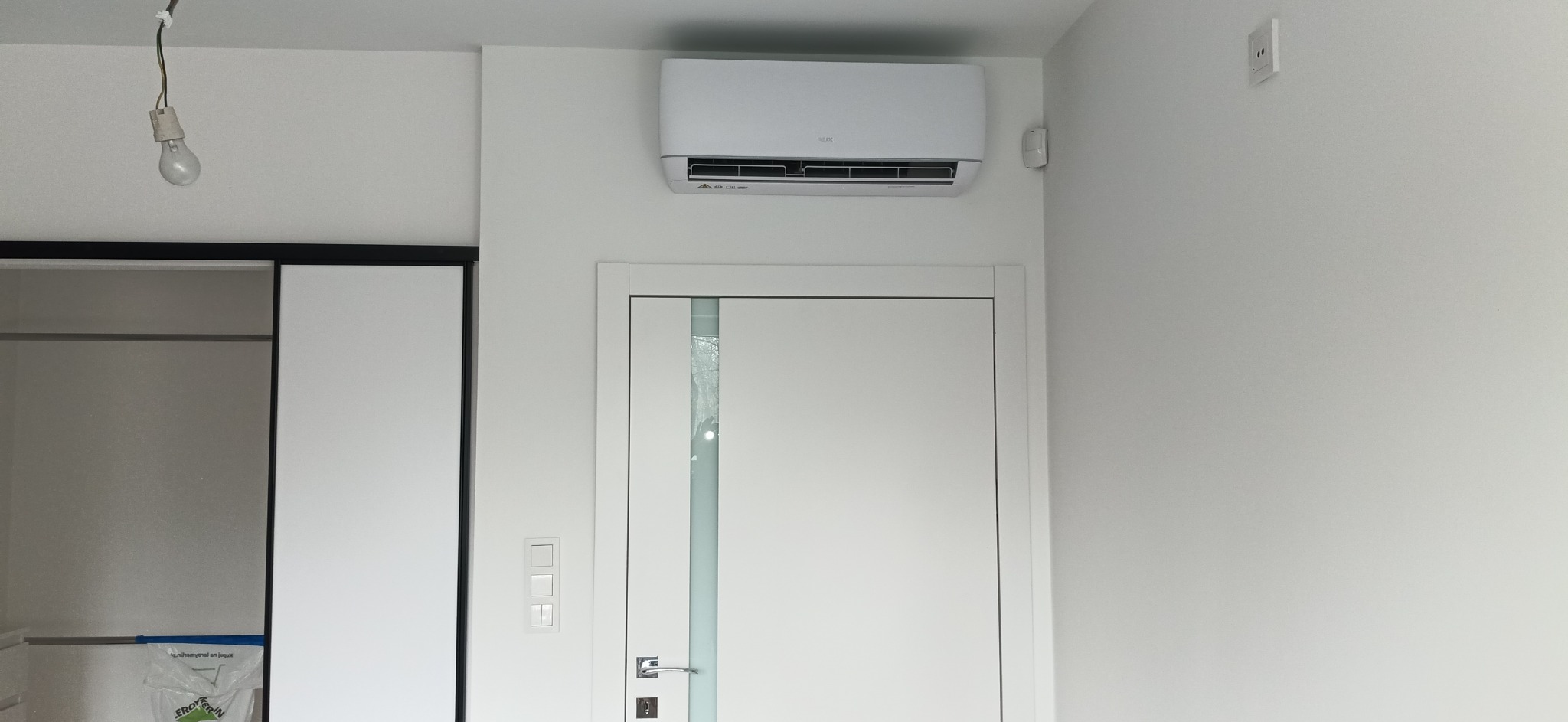 Biała klimatyzacja zamontowana nad białą, nowoczesną ościeżnicą drzwiową w minimalistycznym wnętrzu, widoczny czujnik ruchu obok klimatyzatora oraz wisząca żarówka bez klosza.