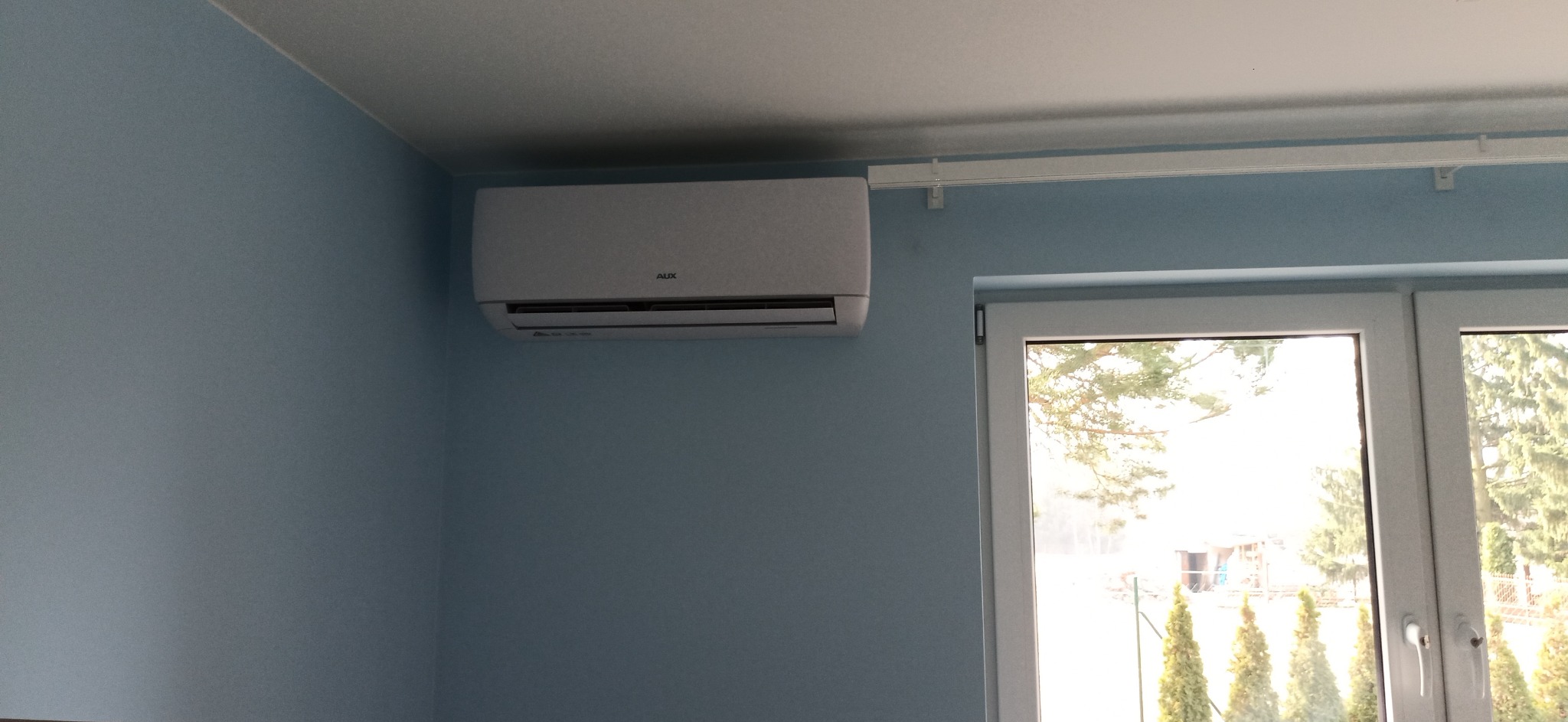 Biały klimatyzator marki AUX zamontowany na błękitnej ścianie w pokoju z widokiem na ogród przez okno.