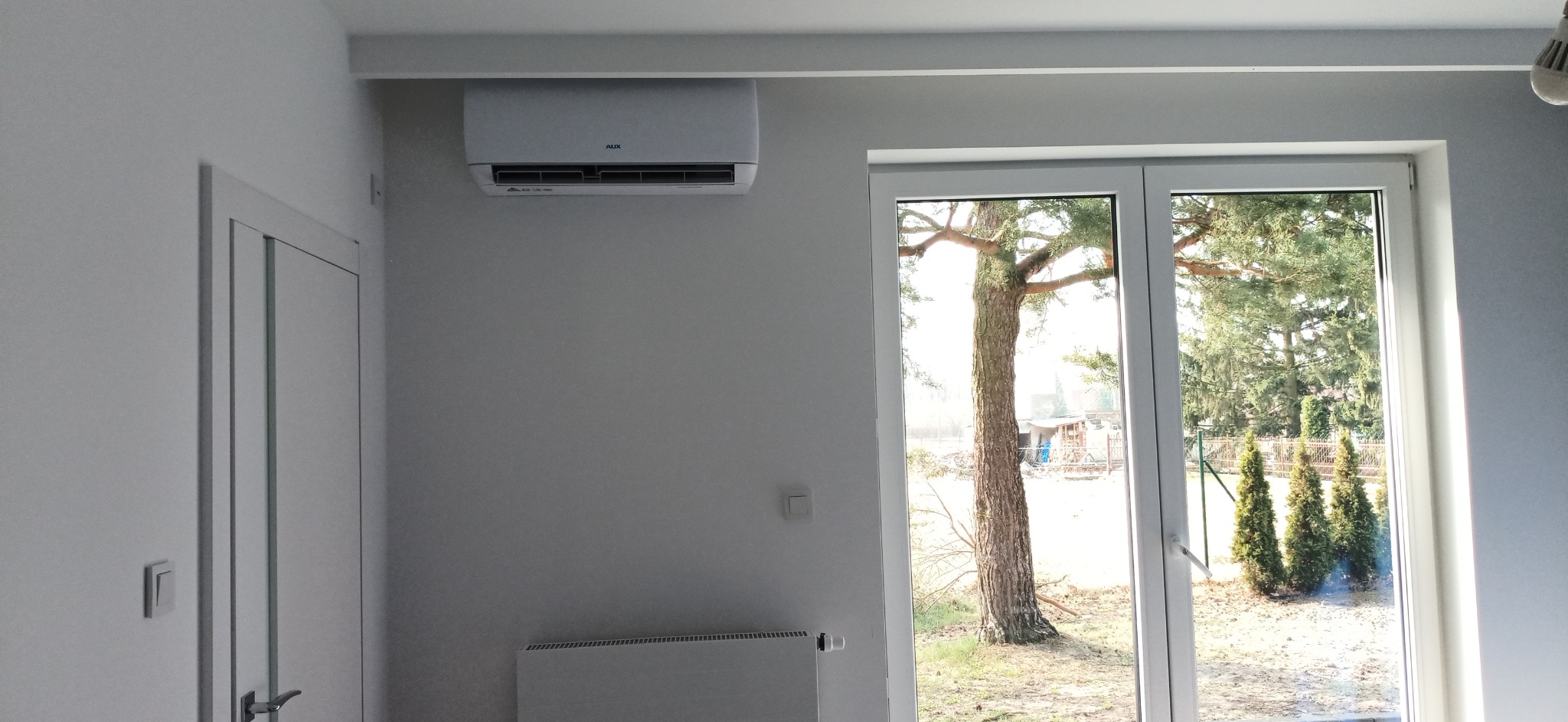 Klimatyzator marki AUX zamontowany w jasnym pokoju w Niedźwiedziu, widok na ogród przez okno balkonowe.