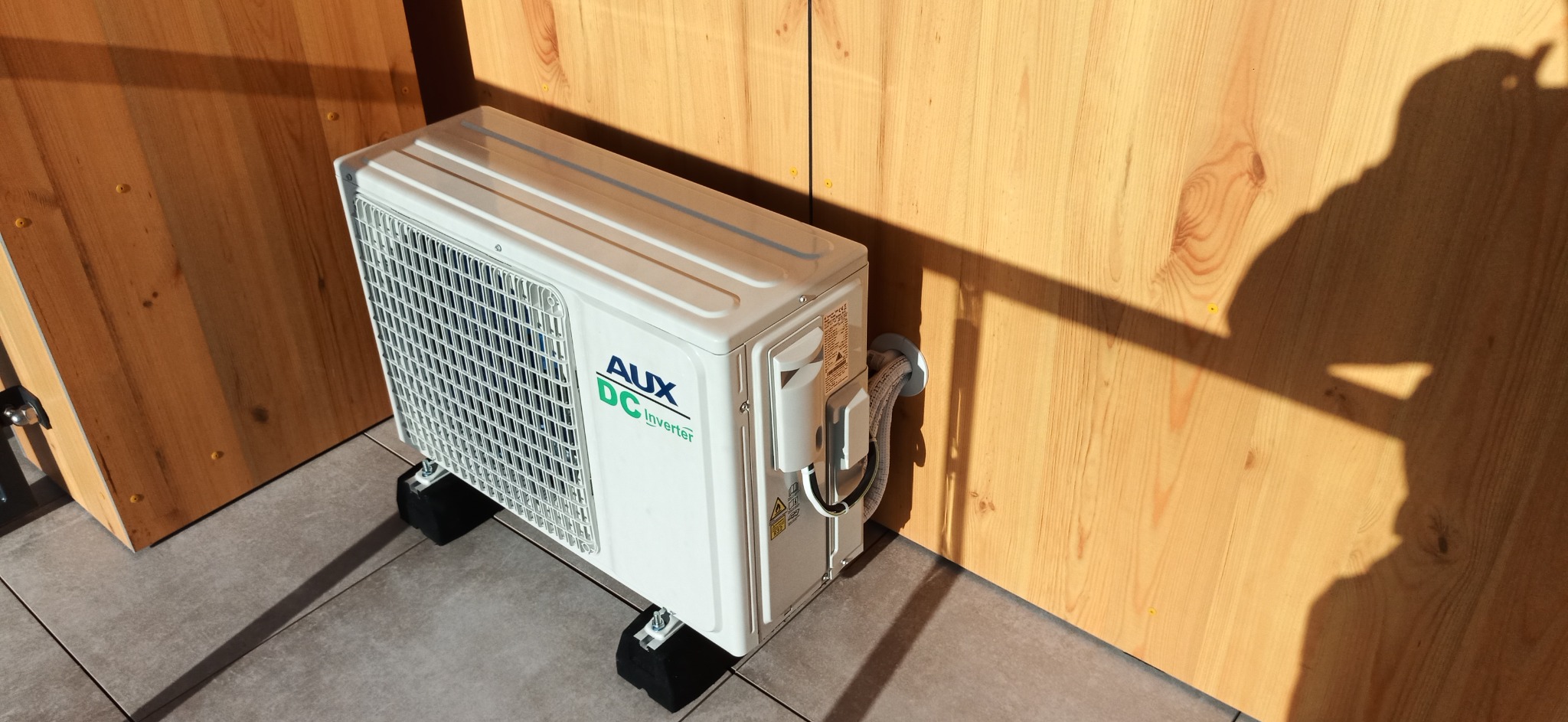 Biała jednostka zewnętrzna klimatyzacji AUX DC Inverter, zamontowana na czarnych podkładkach antywibracyjnych, z widocznymi przewodami przechodzącymi przez ścianę z jasnego drewna, cień osoby...