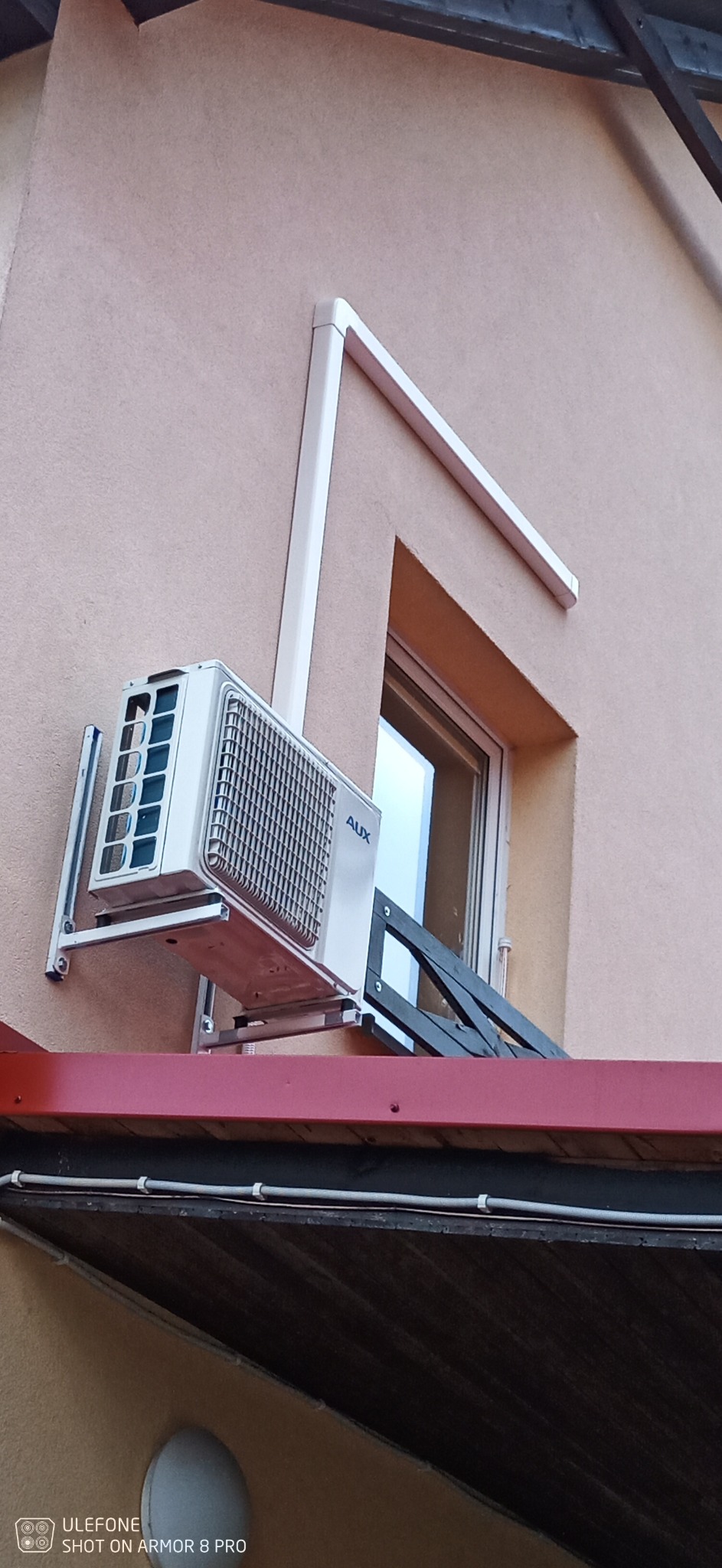 Zewnętrzna jednostka klimatyzacji marki AUX zamontowana na ścianie budynku o jasnej elewacji, widoczne czarne rury odprowadzające skropliny, montaż pod oknem.