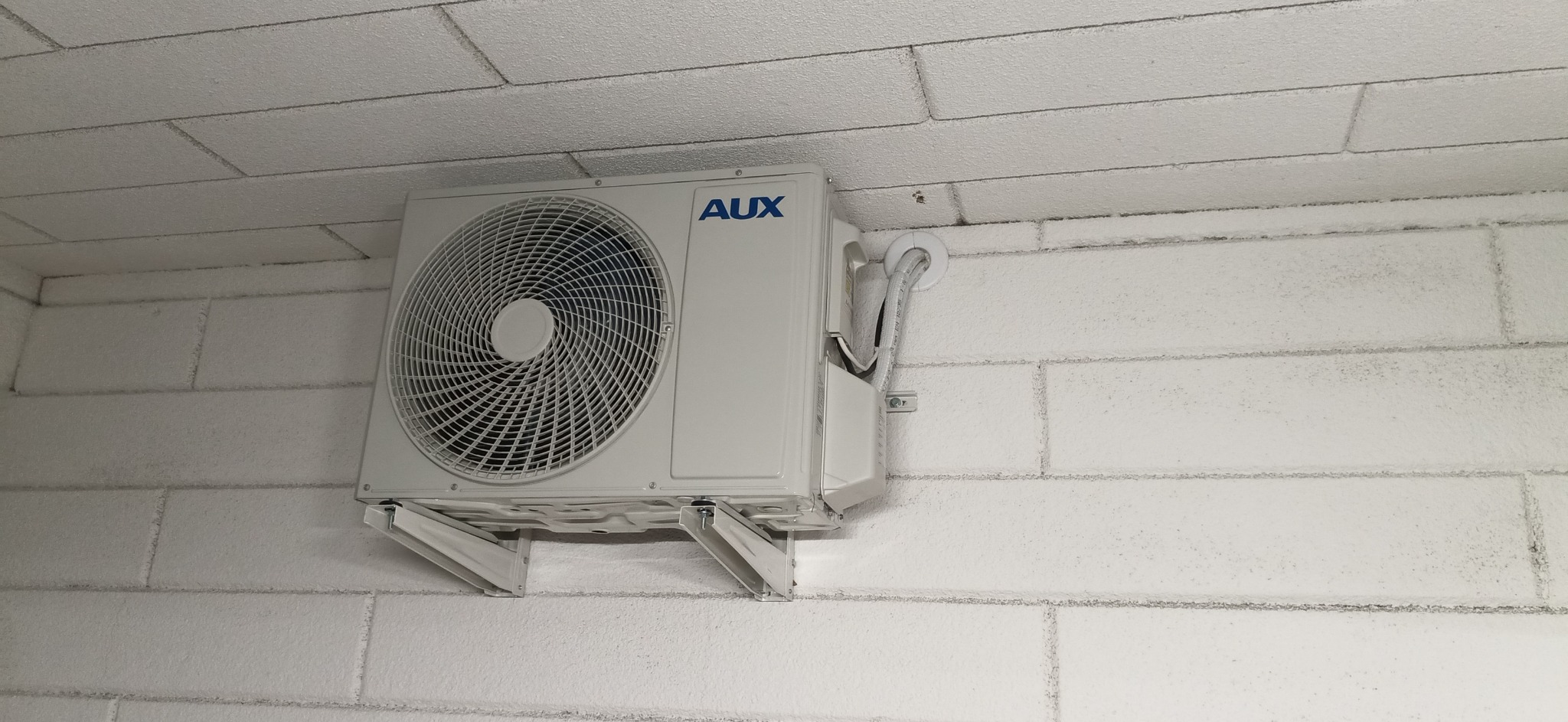 Biała jednostka zewnętrzna klimatyzacji marki AUX zamontowana na białej, ceglanej ścianie wewnętrznej, widoczne przewody w białej maskownicy.