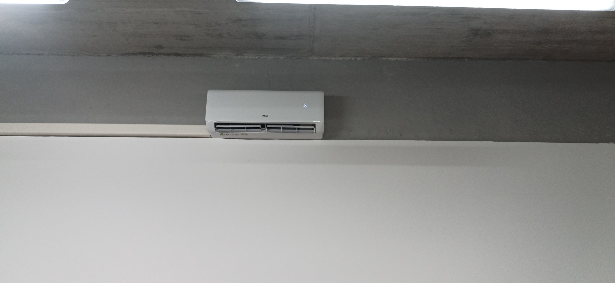 Biały klimatyzator AUX zamontowany na szarej ścianie, widoczny wyświetlacz wskazujący 16 stopni Celsjusza, pod betonowym sufitem.