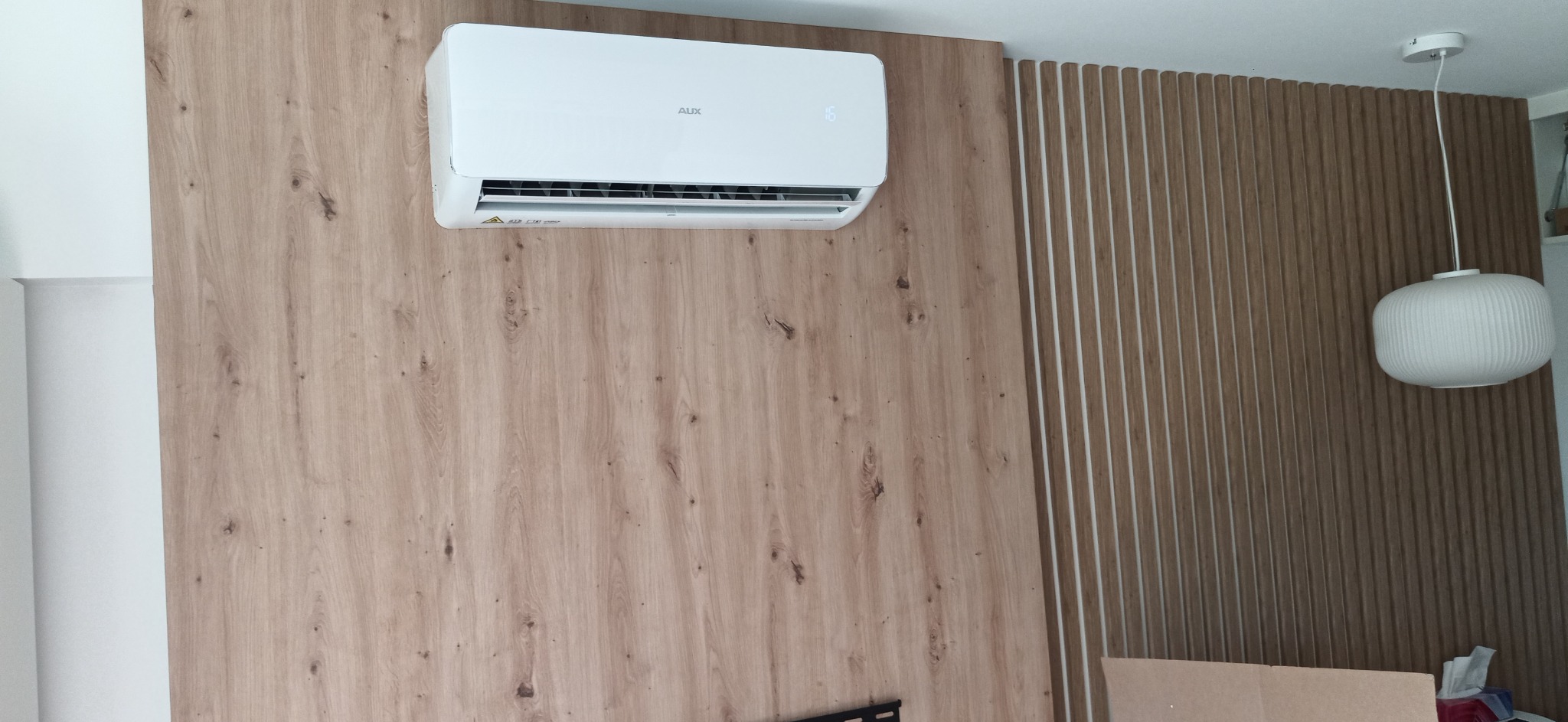 Biały klimatyzator AUX zamontowany na ścianie z panelami drewnopodobnymi, obok dekoracyjne lamele pionowe i wisząca lampa.