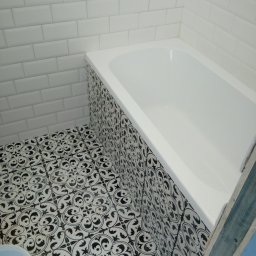 Sebastian Cymba - Wykończona łazienka z białą wanną obudowaną płytkami w czarno-białe wzory, białe kafelki na ścianach ułożone w cegiełkę i wzorzysta podłoga z widocznym fragmentem niebieskiej ściany