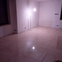 Ułożone płytki podłogowe w jasnym odcieniu beżu, widoczne ślady po pracach wykończeniowych, na ścianach gniazdka elektryczne, w tle lampa podłogowa i okno.