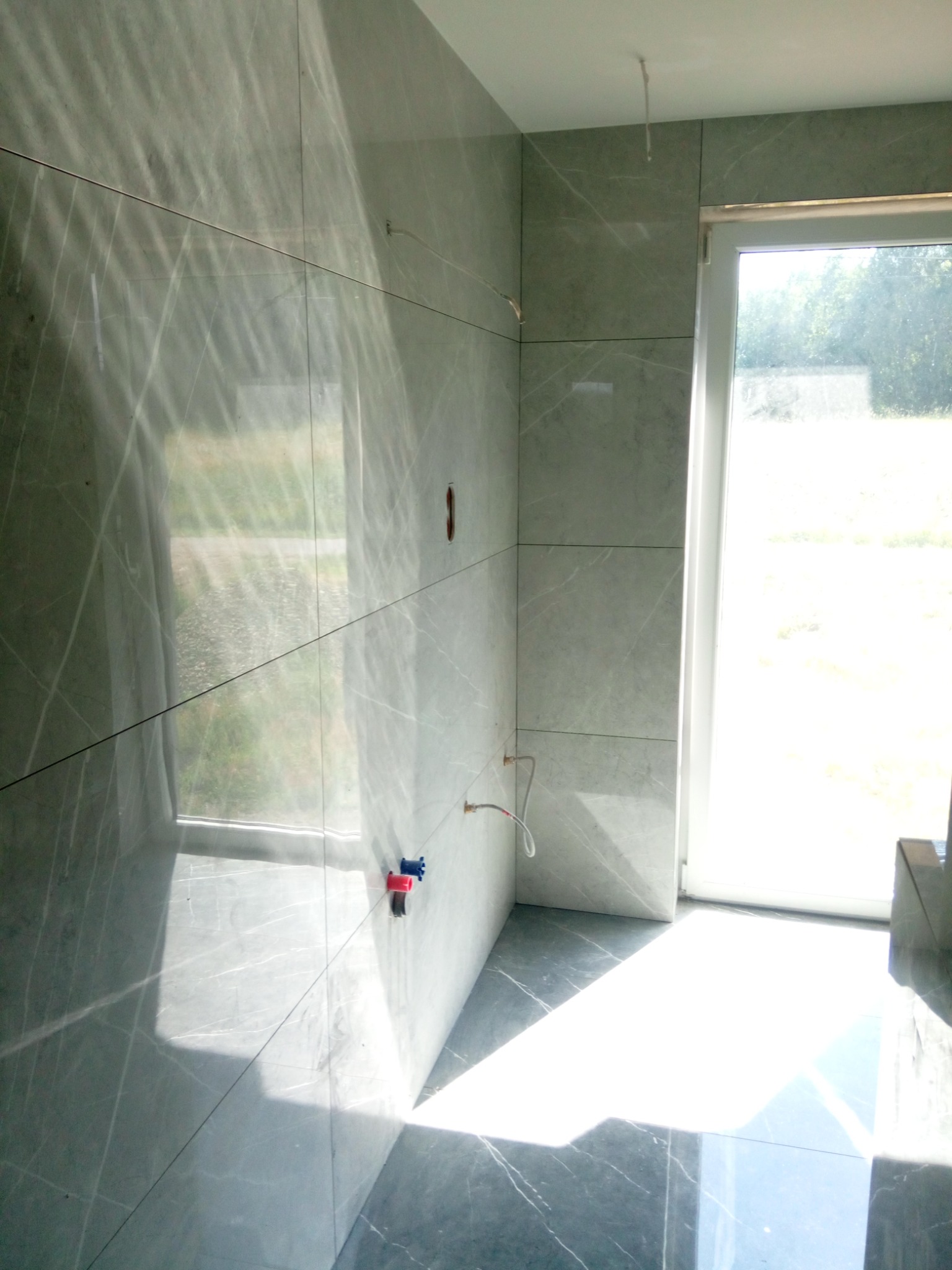 Świeżo ułożone, duże, szare płytki imitujące marmur na ścianach i podłodze łazienki w trakcie wykańczania, z widocznymi wystającymi rurami i przewodami oraz intensywnym światłem słonecznym...