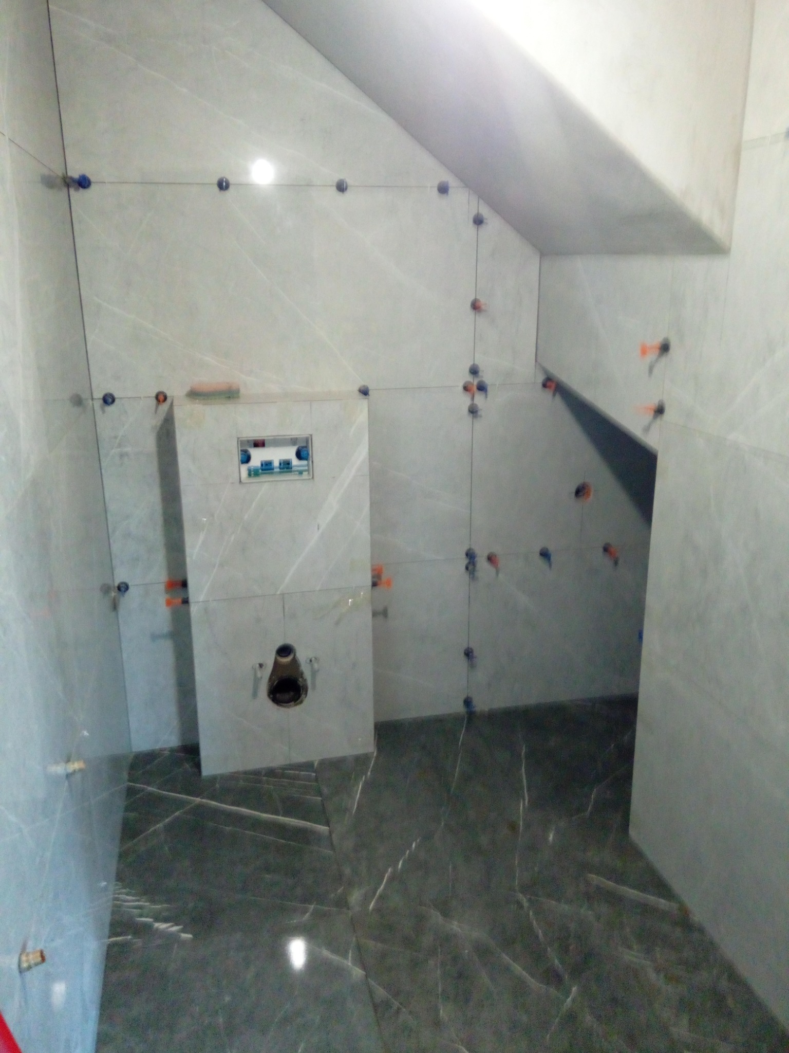 Montaż płytek imitujących marmur w łazience w trakcie realizacji, widoczne krzyżyki dystansowe i stelaż podtynkowy WC ze zbiornikiem, podłoga w ciemniejszym odcieniu.