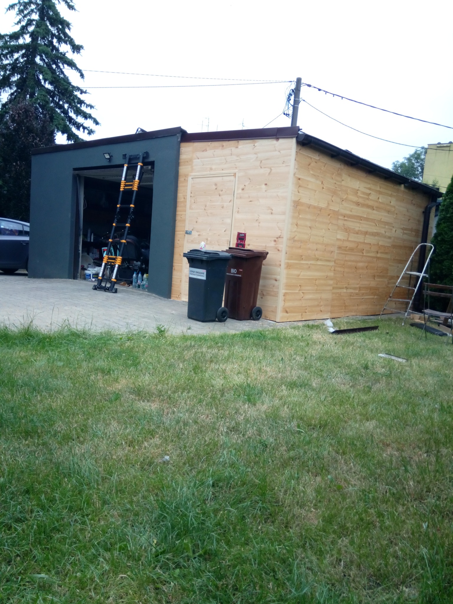 Zewnętrzny widok nowo wybudowanego garażu z drewnianą elewacją i otwartą bramą, przy której oparta jest rozłożona drabina teleskopowa; obok stoją dwa pojemniki na śmieci.