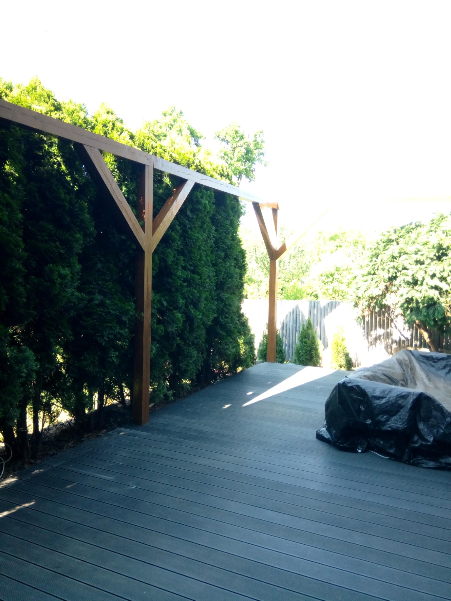 Nowoczesny taras z kompozytowych desek w odcieniach szarości, z drewnianą pergolą i żywopłotem z tui, częściowo zakryty plandeką.
