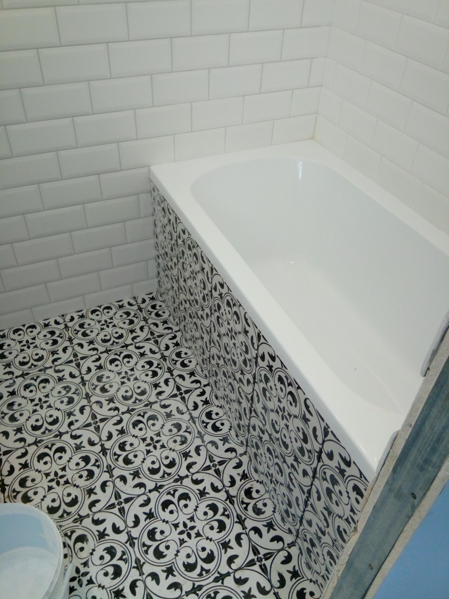 Wykończona łazienka z białą wanną obudowaną płytkami w czarno-białe wzory, białe kafelki na ścianach ułożone w cegiełkę i wzorzysta podłoga z widocznym fragmentem niebieskiej ściany