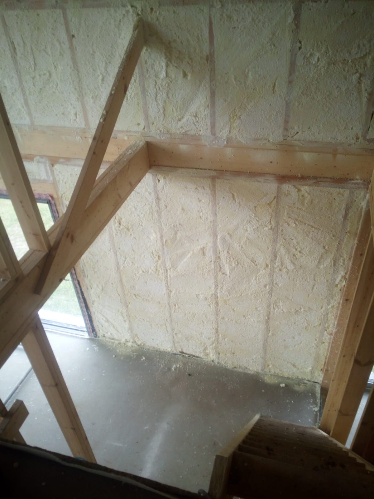 Poddasze w trakcie ocieplania pianką, widoczne drewniane krokwie i świeżo nałożona warstwa izolacyjna o nieregularnej fakturze, okno w tle.