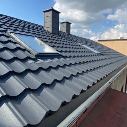 Markowe Dachy - Grafitowy dach z blachy trapezowej z dwoma oknami dachowymi i kominami na tle błękitnego nieba z chmurami. Widoczne rynny.