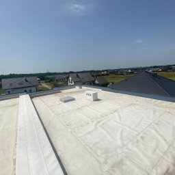 Markowe Dachy - Biała membrana na płaskim dachu budynku jednorodzinnego, widok z góry na tle zabudowy i błękitnego nieba. Widoczne detale łączeń i elementy wentylacyjne.