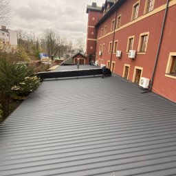 Markowe Dachy - Szary dach z blachy trapezowej na budynku z czerwonej cegły. Widoczny fragment dachu sąsiedniego budynku i pracownik na dachu. Pochmurne niebo w tle.
