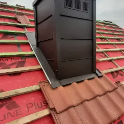 Nowo wybudowany komin wentylacyjny z brązowej blachy na dachu pokrytym czerwoną dachówką ceramiczną, widoczne membrany dachowe i drewniane łaty.