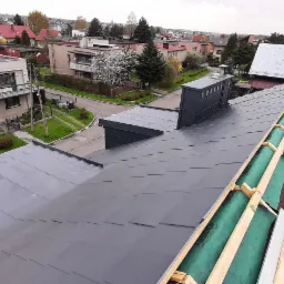 Szary dach z blachy układany na konstrukcji z łat i kontrłat, widoczny komin i fragmenty starego pokrycia, w tle zabudowa miejska.