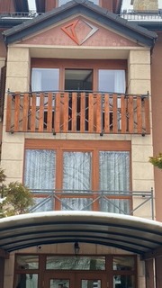 Dwukondygnacyjny budynek z drewnianym balkonem i oknami, widok z dołu, zadaszone wejście, dekoracyjny element w kształcie rombu nad balkonem.