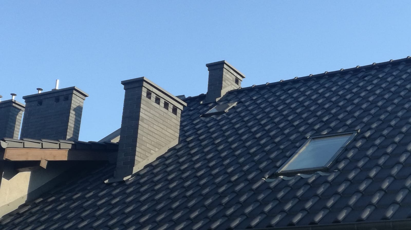 Ciemna dachówka ceramiczna z oknem dachowym i ceglane kominy na tle błękitnego nieba. Widok z dołu, podkreślający solidność wykonania.