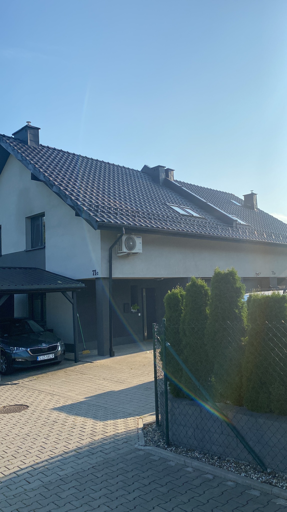 Dom jednorodzinny z ciemną dachówką i widocznymi oknami dachowymi, sfotografowany w słoneczny dzień z poziomu chodnika. Widoczny fragment ogrodzenia i podjazdu.