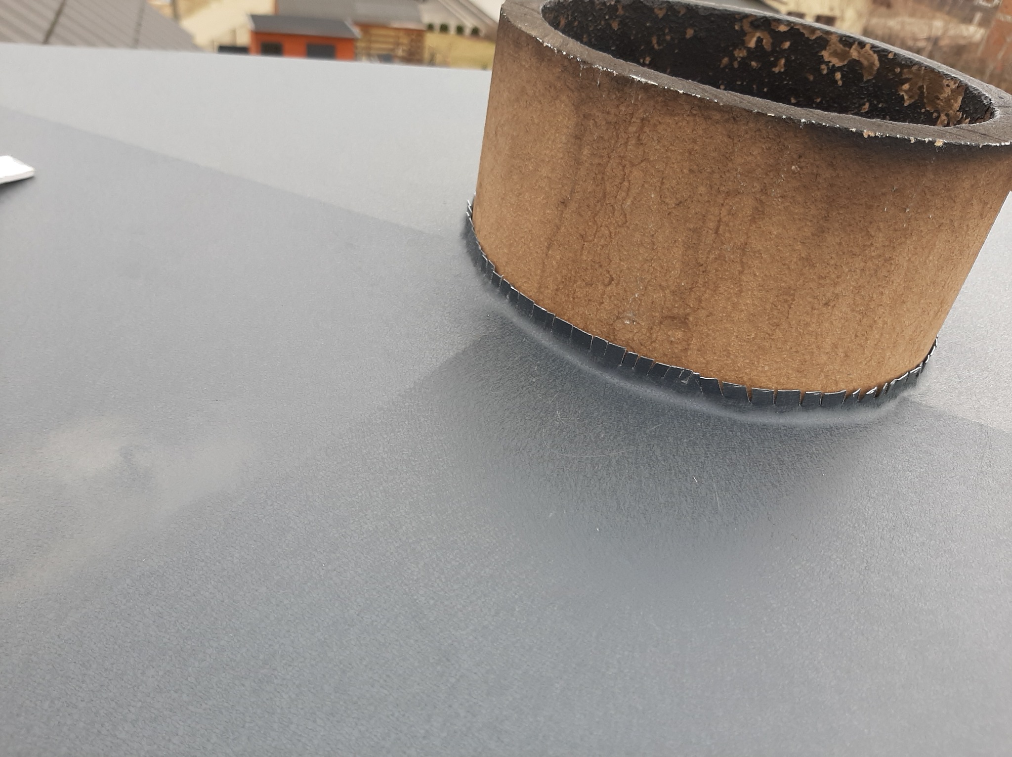 Szary dach z blachy z obróbką komina w postaci brązowego, okrągłego elementu. Widoczne mocowania blacharskie. Detal wykonania.