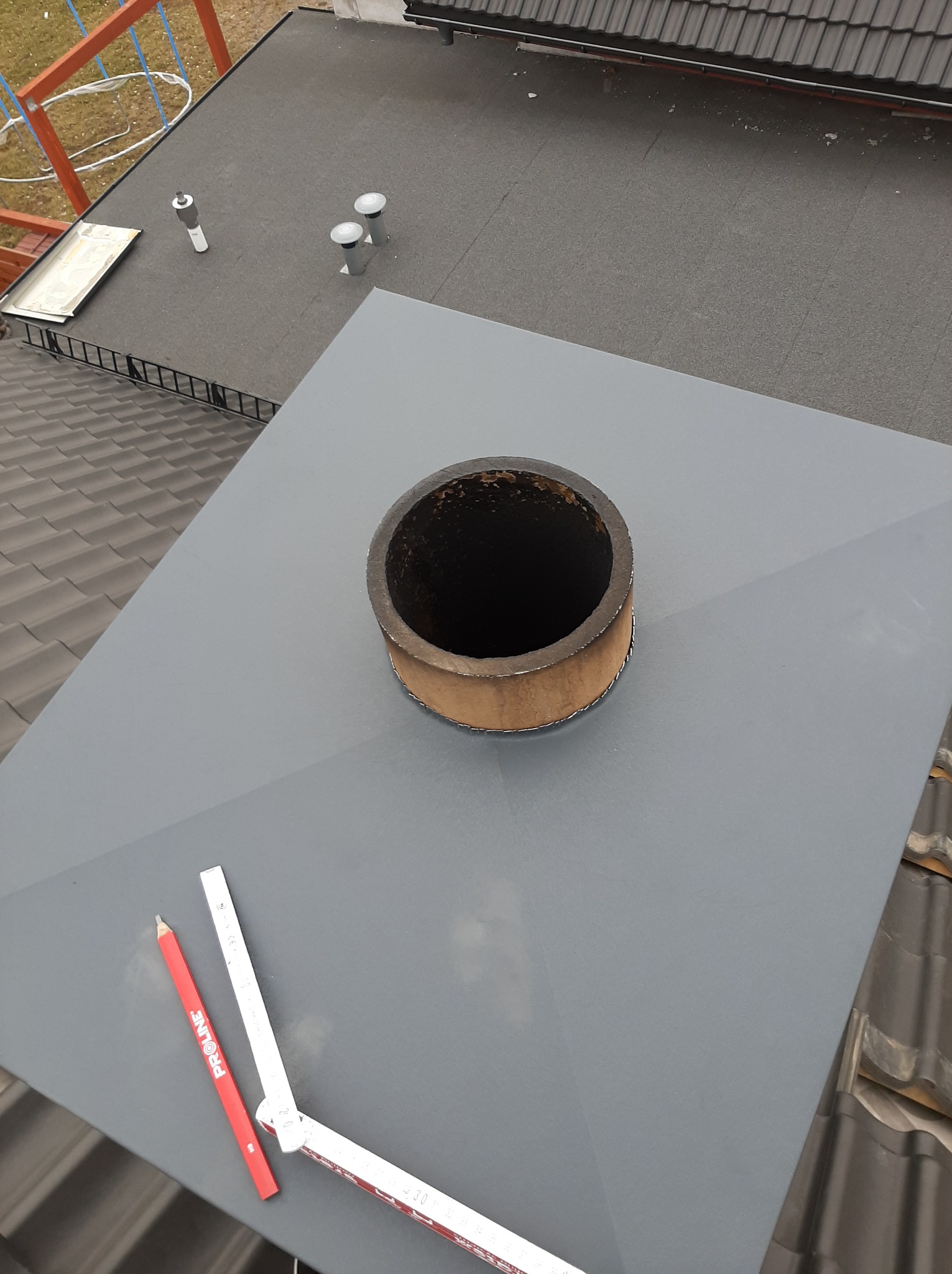 Szary, metalowy komin na dachu z dachówki, obok leżą narzędzia: czerwona kredka i składana miarka. Widok z góry na fragment dachu pokrytego papą.