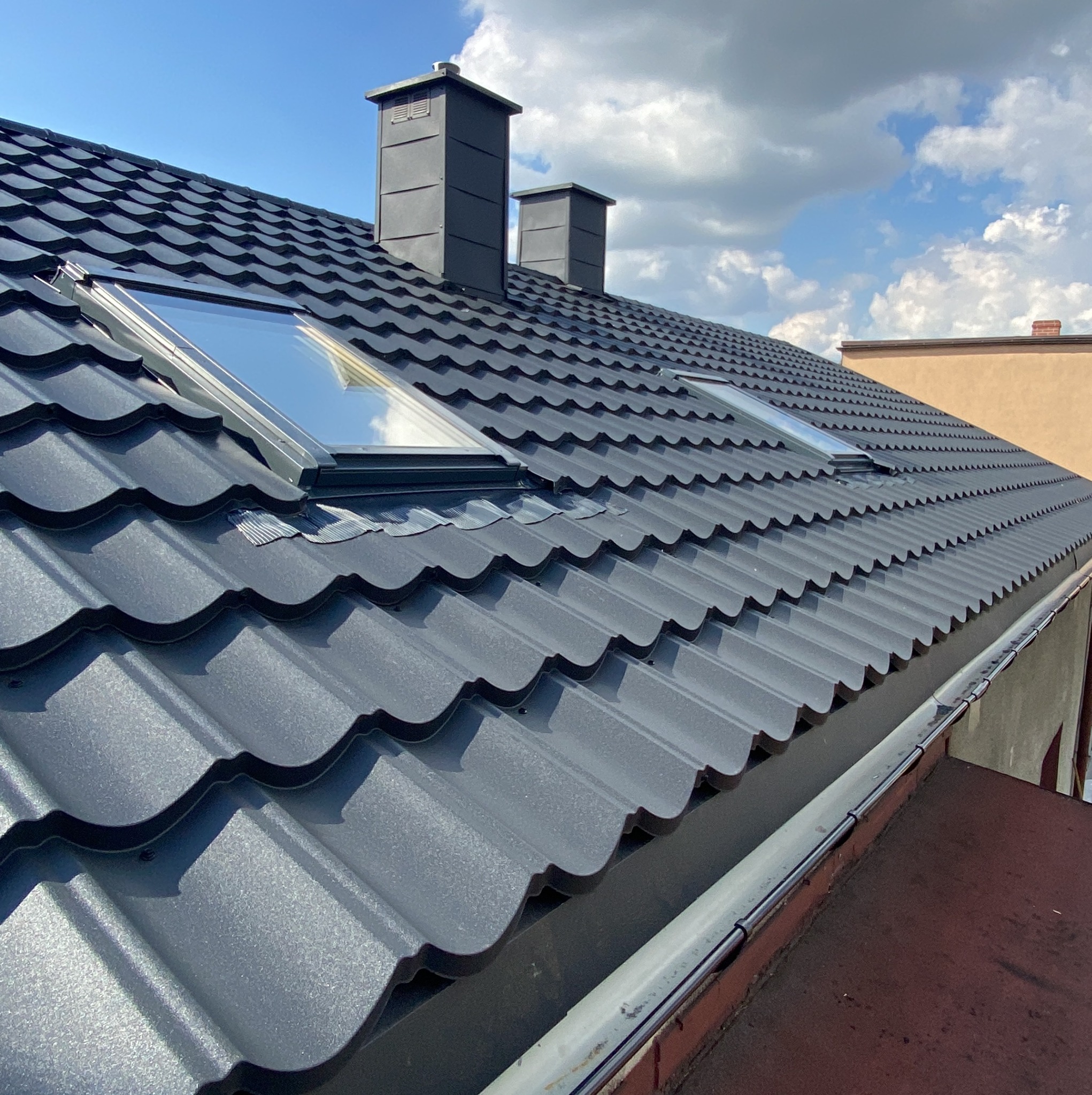 Grafitowy dach z blachy trapezowej z dwoma oknami dachowymi i kominami na tle błękitnego nieba z chmurami. Widoczne rynny.