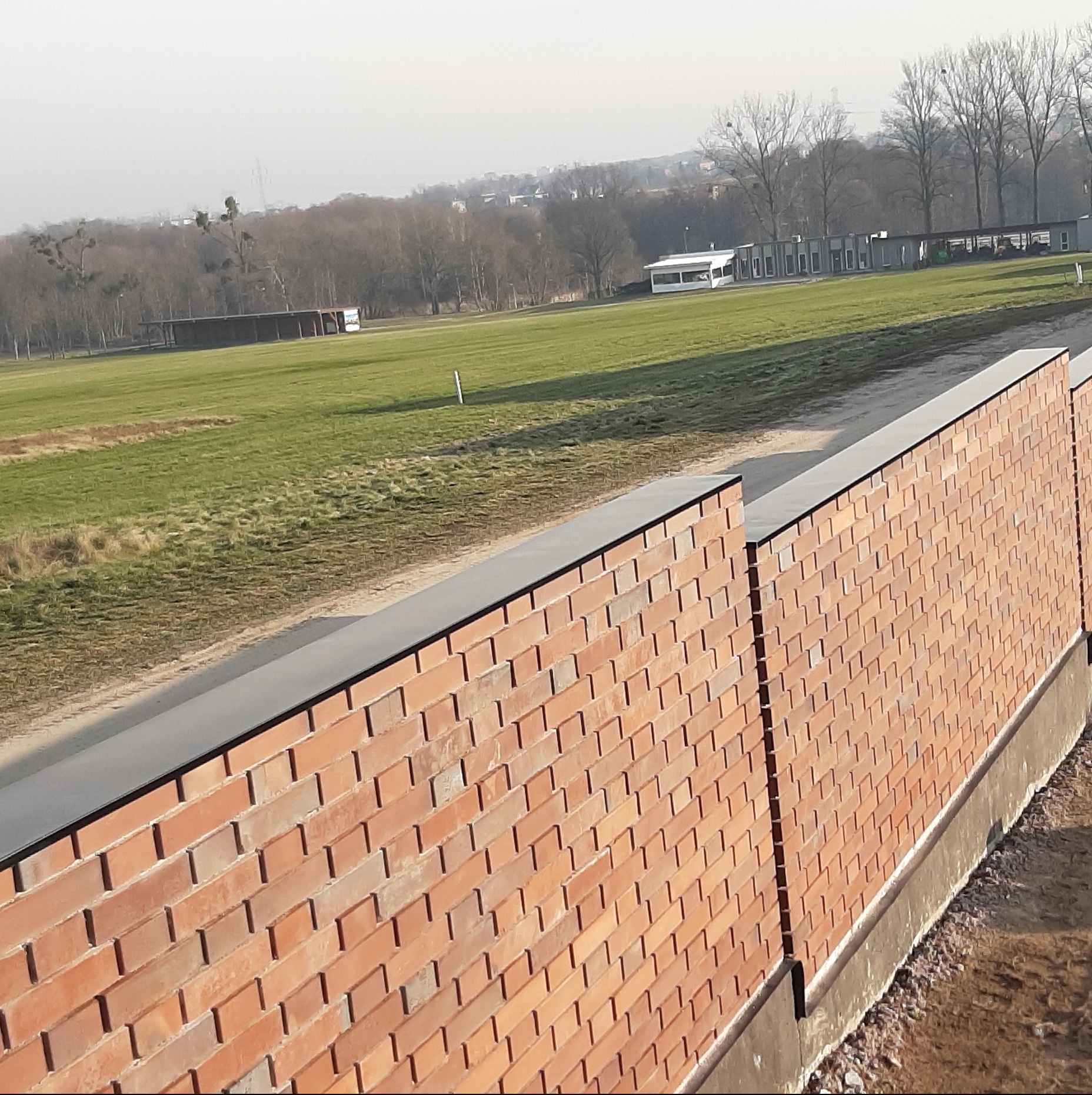Mur z czerwonej cegły z czarnym wykończeniem na tle zielonego pola i budynków w oddali, wykonany pod kątem, ukazujący detale ceglanej konstrukcji.