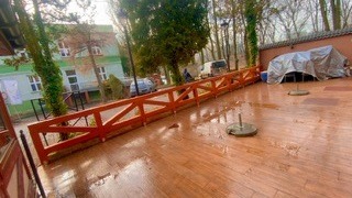 Drewniany taras po deszczu z widoczną balustradą i budynkami w tle. Mokra nawierzchnia odbija światło, tworząc refleksy. Widoczne drzewa i elementy otoczenia.