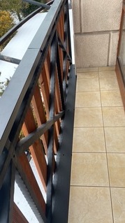 Barierka balkonowa z drewnianymi elementami i czarną metalową konstrukcją, widok z góry na jasne płytki podłogowe i fragment okna. Jesienne drzewo w tle.