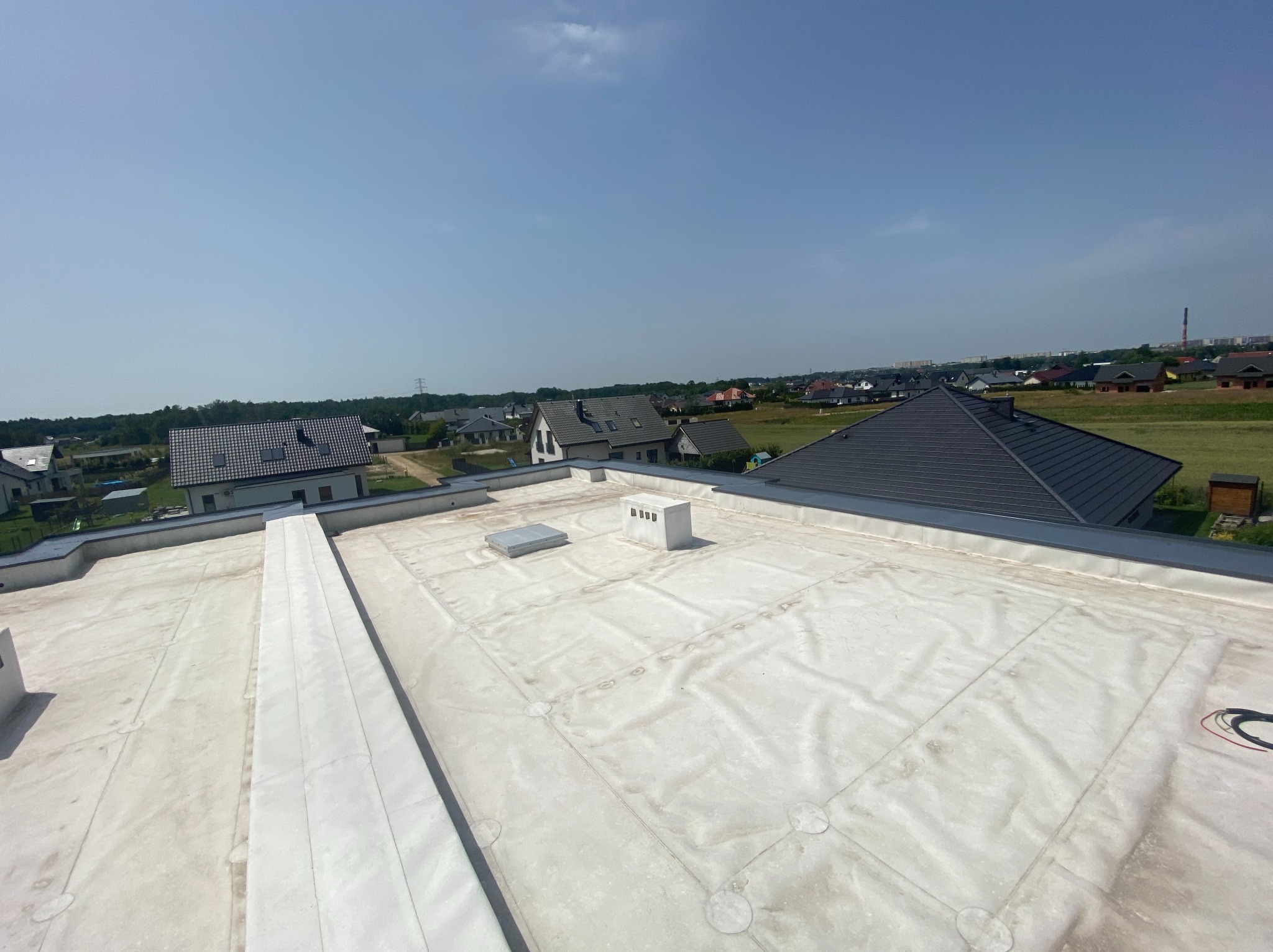 Biała membrana na płaskim dachu budynku jednorodzinnego, widok z góry na tle zabudowy i błękitnego nieba. Widoczne detale łączeń i elementy wentylacyjne.