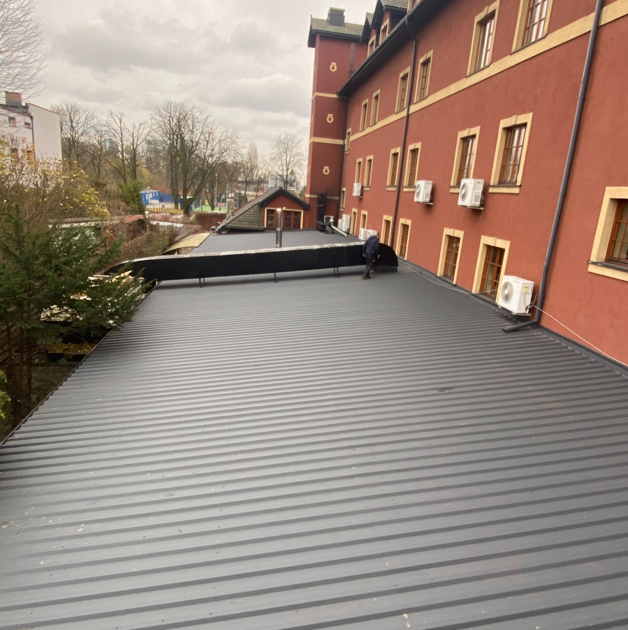 Szary dach z blachy trapezowej na budynku z czerwonej cegły. Widoczny fragment dachu sąsiedniego budynku i pracownik na dachu. Pochmurne niebo w tle.