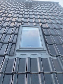 Czarne dachówki ceramiczne z oknem połaciowym i obróbką blacharską, widok z dołu pod kątem, pochmurne niebo odbija się w szybie okna.