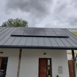 Panele fotowoltaiczne, czarne, zamontowane na szarym, metalowym dachu domu jednorodzinnego z drewnianymi elementami wykończeniowymi w pochmurny dzień.