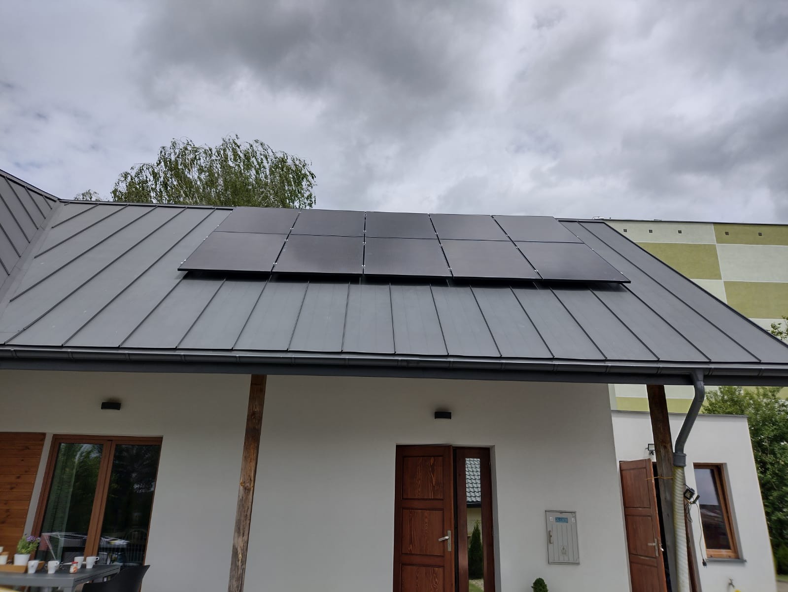 Panele fotowoltaiczne, czarne, zamontowane na szarym, metalowym dachu domu jednorodzinnego z drewnianymi elementami wykończeniowymi w pochmurny dzień.