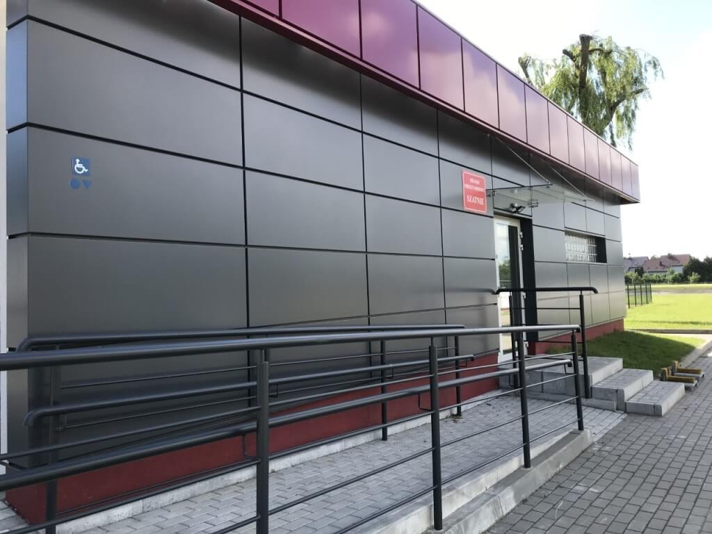 Nowoczesny budynek szatni z panelami w kolorze grafitowym, oznaczony znakiem dla niepełnosprawnych, z podjazdem i schodami z kostki brukowej.