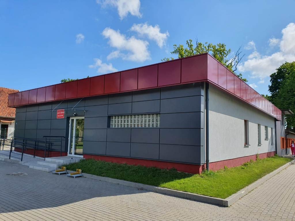 Gotowy budynek parterowy z szarymi panelami elewacyjnymi, czerwonym dachem i rampą dla niepełnosprawnych przy wejściu, otoczony kostką brukową i zielenią.