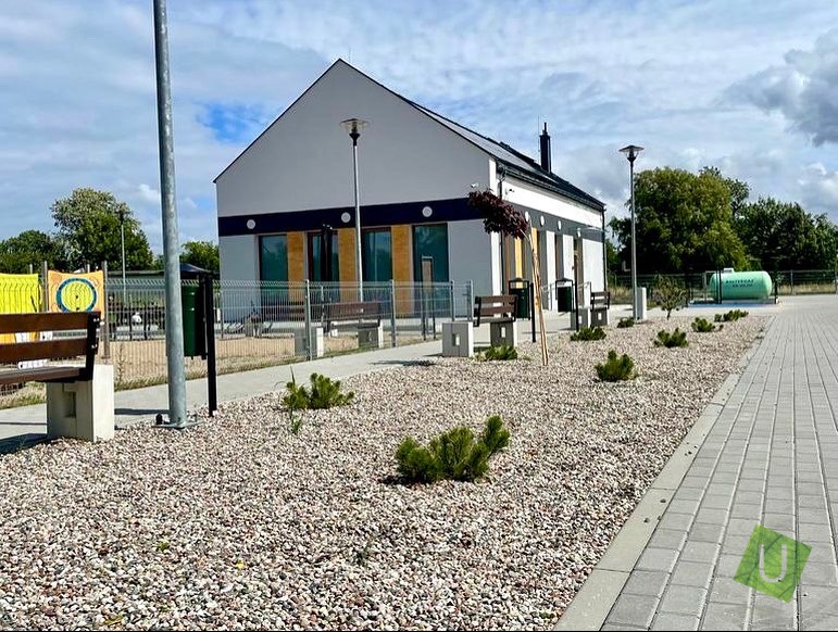 Nowoczesny budynek z jasną elewacją, ciemnym pasem dekoracyjnym i drewnianymi elementami, otoczony żwirowym terenem z nasadzeniami, ławkami i chodnikiem z kostki brukowej. Na drugim planie widoczny...